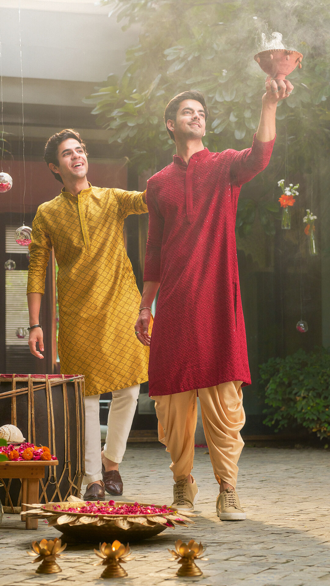 Two young men in traditional kurta pajamas performing Durga Puja rituals with dhunuchi smoke and festive decorations around.